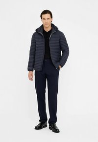 Mereväe paks jope horisontaalse õmblusega ja kinnitatud kapuutsiga, kombineeritud tumedate pükste ja mustade kingadega. Sile tekstuur, minimaalsed aktsendid.