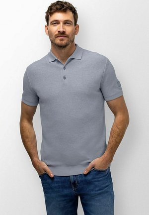 Man met kort bruin haar en baard, die een lichtgrijs poloshirt en blauwe spijkerbroek draagt, staand met handen in de zakken tegen een effen witte achtergrond.