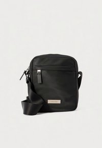 Zwarte rechthoekige stoffen crossbodytas met een voorvak met ritssluiting, een verstelbare riem en een zilverkleurig logoplaatje aan de voorkant.