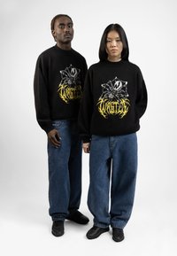 Fekete, oversized pulóverek grafikai nyomattal, lazán illeszkedő kék farmernadrággal párosítva. Mindkettő fekete sportcipőt visel.