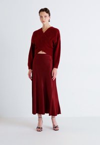 Rød innpakket genser laget av teksturert stoff, parret med et matchende slim-fit skjørt. Modellen har på seg svarte og hvite mønstrede høyhælte sandaler.