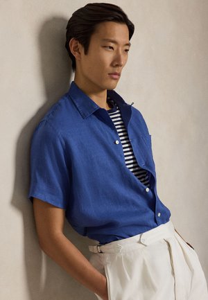 Fiatal férfi, aki nekidől a falnak, kék rövid ujjú inget visel csíkos póló felett és fehér, magas derekú nadrágot, kezét a zsebében tartva.