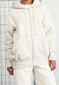 Ljusbeige zip-up hoodie i mjukt tyg, med en rymlig huva, ärmar och två framfickor. Litet vitt logotyp på bröstet.