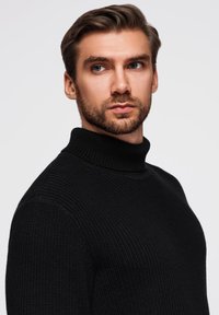 Czarny dzianinowy sweter z wysokim kołnierzem o teksturowanej tkaninie, dopasowanej sylwetce, noszony przez modela o jasnej karnacji i brodzie.