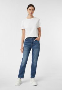 Weißes Baumwoll-T-Shirt mit kurzen Ärmeln, kombiniert mit blauen geraden Jeans aus Denim. Das Model trägt weiße Sneakers und steht mit den Händen in den Taschen.