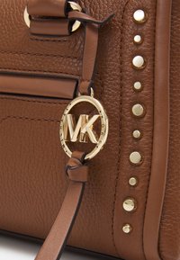 Brązowa torebka skórzana o teksturowanej powierzchni z pozłacanymi wkrętami i okrągłym breloczkiem z logo Michael Kors zawieszonym na skórzanym pasku.
