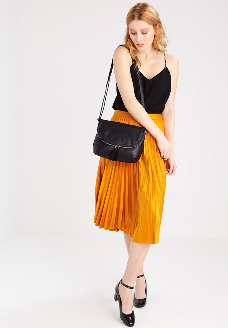 Bolso de cuero negro con diseño de solapa y cierre de cremallera, llevado al hombro. La modelo viste una blusa negra y una falda midi amarilla con plisados.
