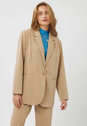 Blazer beige en tissu léger avec une fermeture à un bouton, des revers crantés et deux poches à l'avant ; porté par-dessus un chemisier bleu à motifs.