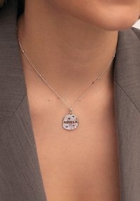 Collier en argent avec un pendentif rond gravé "ABUELA" décoré de petites pierres précieuses colorées, porté par une personne en blazer gris.