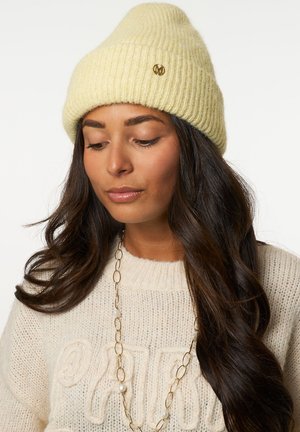 Vrouw met lang donker haar, gekleed in een crèmekleurige gebreide trui, een gouden ketting met parels en een lichtgele geribbelde gebreide muts.
