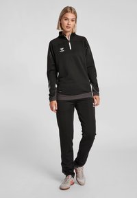 Sort atletisk zip-up top med grå detaljer, teksturerede ærmer og logo. Kombineret med matchende sorte bukser og lyse træningssko.