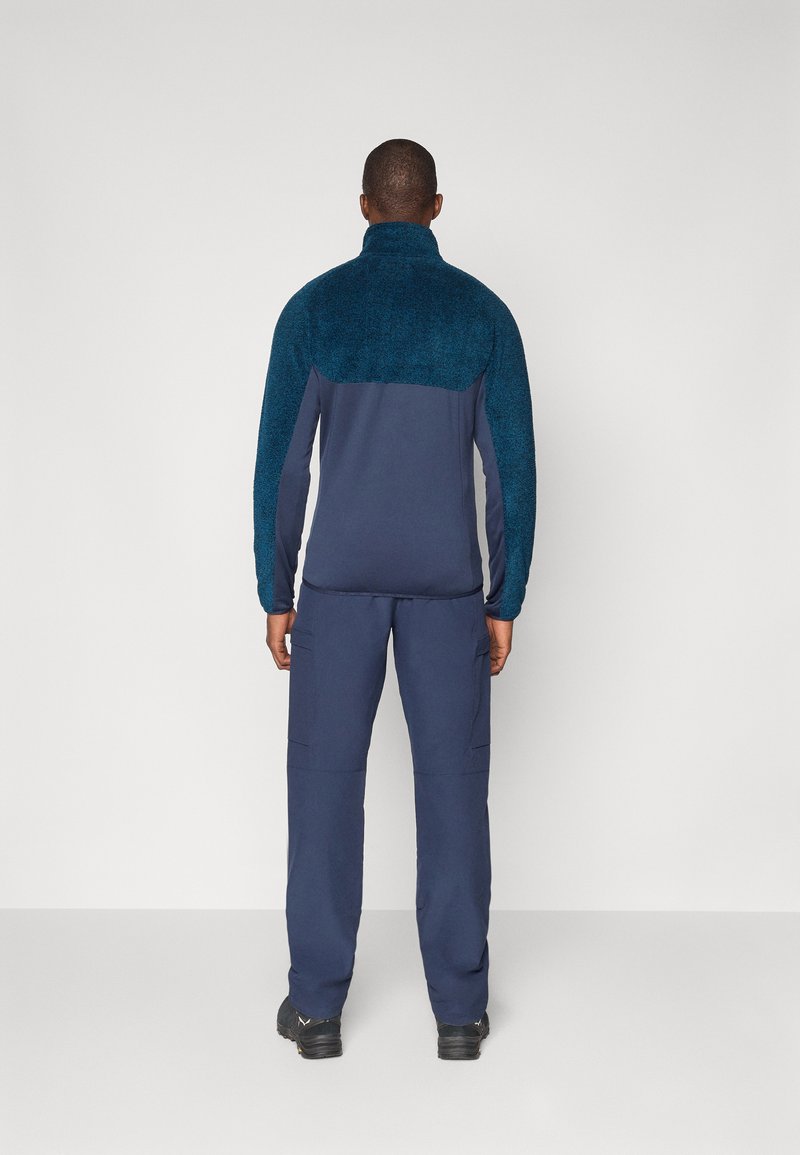 Granatowy sweter z puszystą, turkusową górną częścią, dopasowany do granatowych spodni. Sweter ma wysoki kołnierz i teksturowany materiał.