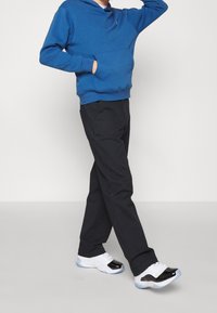 Felpa blu con tasca frontale, pantaloni neri e sneaker bianche con accenti neri e suole blu. Texture liscia, vestibilità casual.