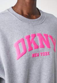 Grå fleecetröja med stora rosa blockbokstäver som säger "DKNY" och "NEW YORK." Rund hals med ribbad struktur på kragen.