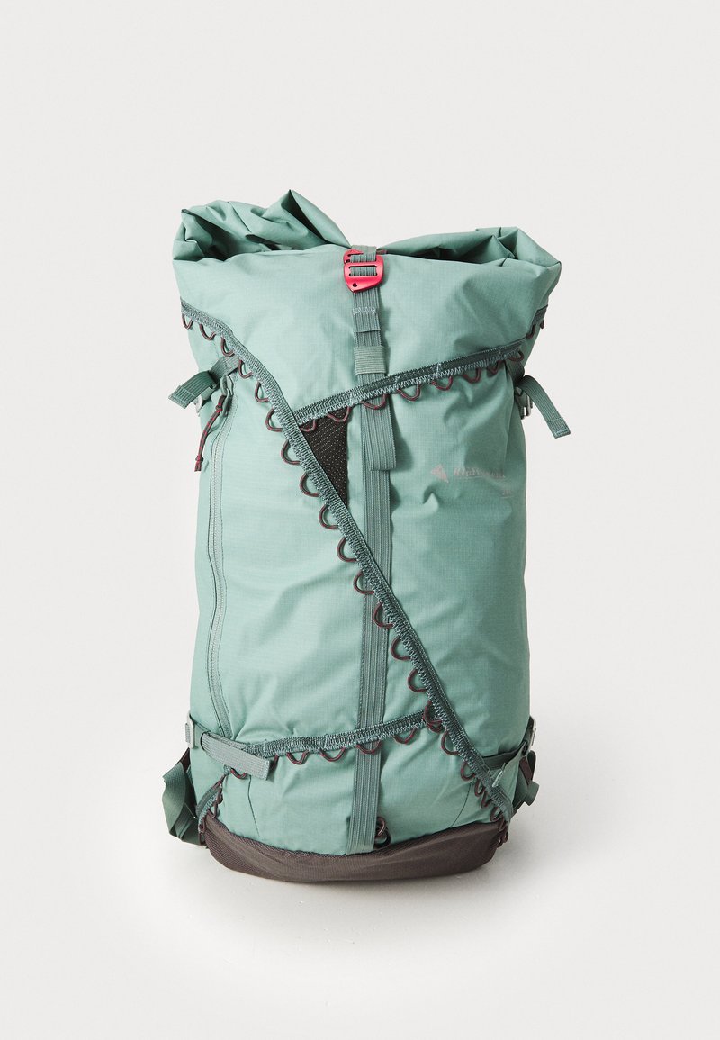 Sac à dos vert à rabat avec une surface texturée, équipé de quincaillerie rose et d'une poche latérale en mesh. Accent de piqûres festonnées décoratives.