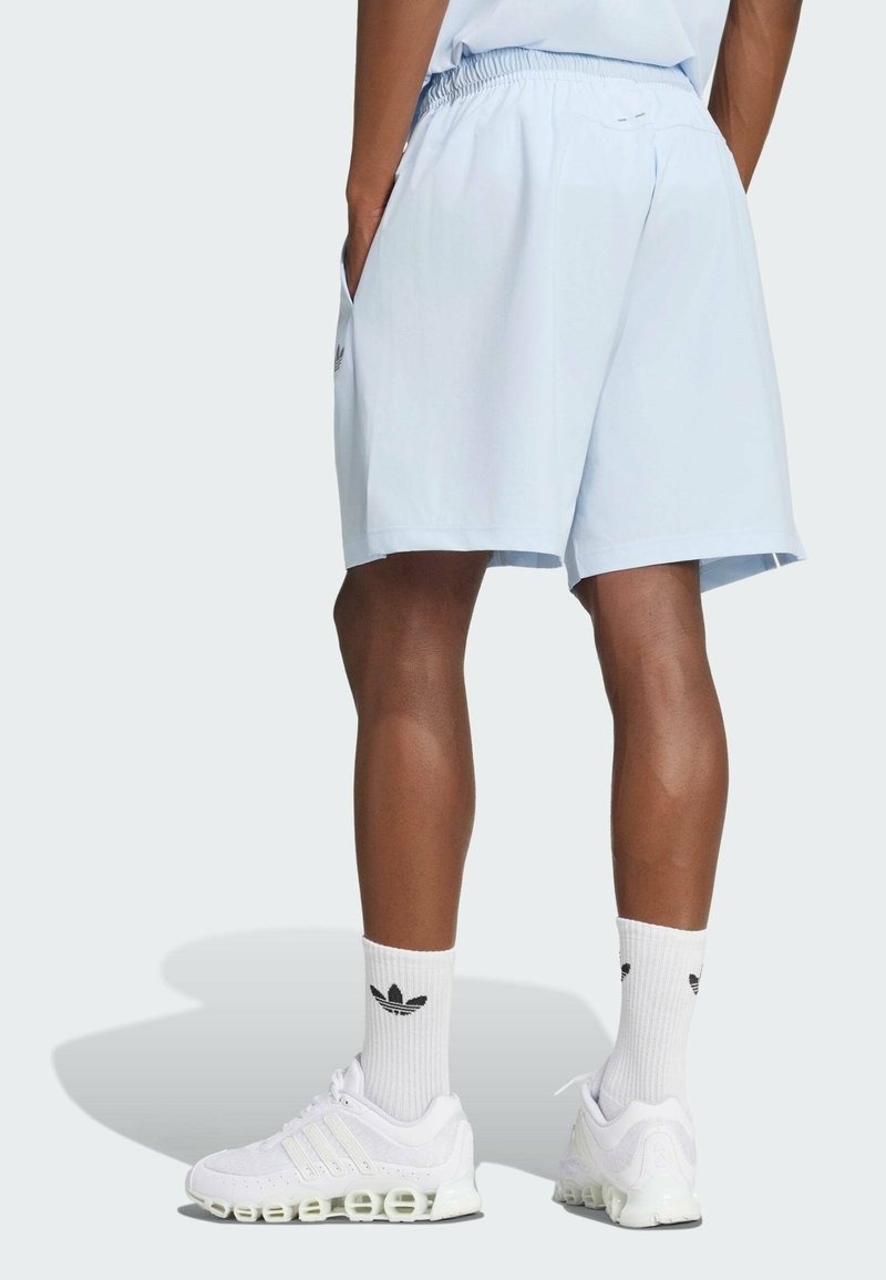 Persona con pantalones cortos azul claro, zapatillas blancas Adidas con suela única y calcetines blancos con el logo de Adidas, de pie con las manos en los bolsillos.