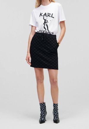 Femme portant un t-shirt blanc à motif avec impression de guitariste, jupe noire à motifs et bottines noires avec accents blancs, debout devant un fond uni.