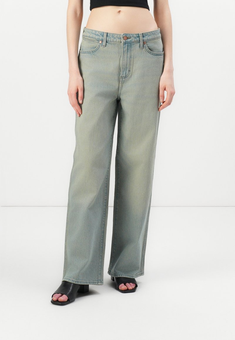 Personne portant un jean bleu délavé taille haute, jambes larges, et des sandales noires à talons ouvertes, debout devant un fond blanc.