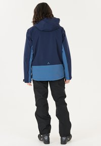 Waterdichte jas met een donkerblauwe bovenkant en een lichtblauwe onderkant, voorzien van een capuchon en zichtbare merkopdruk. Zwarte broek maakt de outfit compleet.