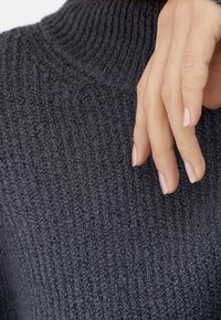 Dunkelgrauer Rollkragenpullover mit strukturiertem Rippenmuster; Hand ruht auf dem Stoff und zeigt glatte, natürliche Nägel.