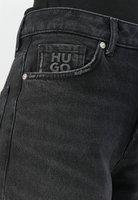 Black denim jeans close-up showing front pocket with stitched small pocket featuring "HUGO" branding and metal rivets.