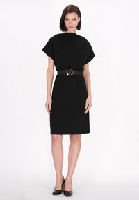 Robe noire avec un design ample et des manches chauve-souris, dotée d'une taille cintrée et d'une ceinture en boucle géométrique. Texture lisse ; longueur au genou. Talons noirs.