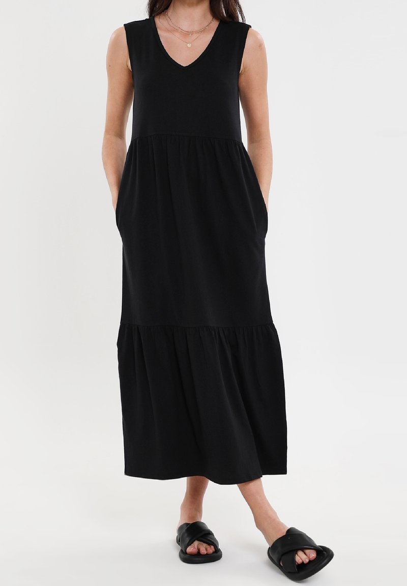Femme portant une robe midi noire sans manches avec poches et des sandales noires à enfiler, debout devant un fond blanc.