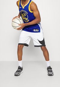 Maglia da basket blu e gialla con il logo dei "Golden State Warriors", pantaloni corti bianchi con accenti neri, tiene in mano un pallone da basket, indossa scarpe da ginnastica grigie.