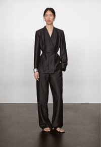 Schwarze, strukturierte Wickeljacke mit Taillengehängter, kombiniert mit passenden, weit geschnittenen Hosen und minimalistischen Sandalen, die eine klare, moderne Silhouette bietet.