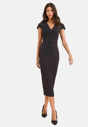 Frau trägt ein figurbetontes schwarzes Midikleid mit kurzen Kapuzenärmeln, V-Ausschnitt, tailliertem Gürtel und schwarzen spitzen High Heels, steht vor einem schlichten Hintergrund.