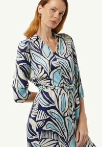 Blaues und cremefarbenes Kleid mit einem Blattmuster, einem Kragen, dreiviertelÄrmeln und einem gebundenen Gürtel in der Taille. Glatte Stofftextur.