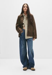 Hnědá semišová bunda přes károvanou košili. Široké modré džíny a hnědé semišové boty. Hladká textura, uvolněný střih, výrazné knoflíky na bundě.