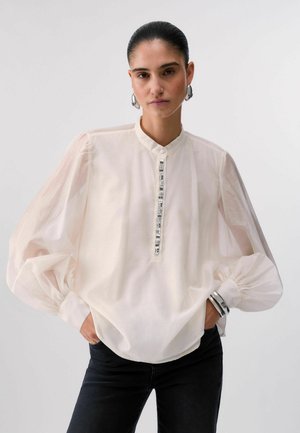 Donna con capelli raccolti all’indietro indossa una camicetta bianca trasparente con maniche a sbuffo, dettaglio gioiello sul davanti, orecchini e braccialetti d'argento, posando con le mani nelle tasche.