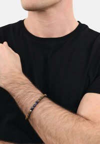 Braccialetto di perline con pietre marroni, gialle e blu su un cinturino flessibile, indossato su un polso su uno sfondo di t-shirt nera.