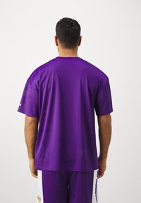 Mężczyzna zwrócony tyłem, ubrany w fioletową sportową koszulkę z siateczki i pasujące spodenki z białymi bocznymi panelami oraz małym logo.