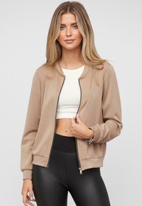 Beige bomberjack met een gladde textuur, geribde manchetten en een ritssluiting aan de voorkant. Te combineren met een witte crop top en zwarte leggings.