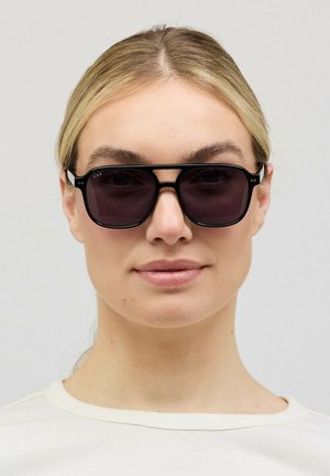 Vrouw met licht haar die zwarte rechthoekige zonnebril en een wit shirt met ronde hals draagt, kijkt recht vooruit tegen een effen achtergrond.