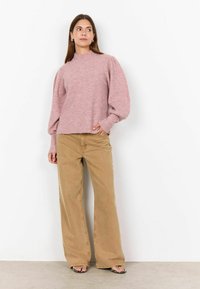 Hellrosa gestrickter Pullover mit hohem Kragen und Puffärmeln, kombiniert mit hellbraunen weit geschnittenen Hosen. Das Model steht vor einem schlichten Hintergrund.