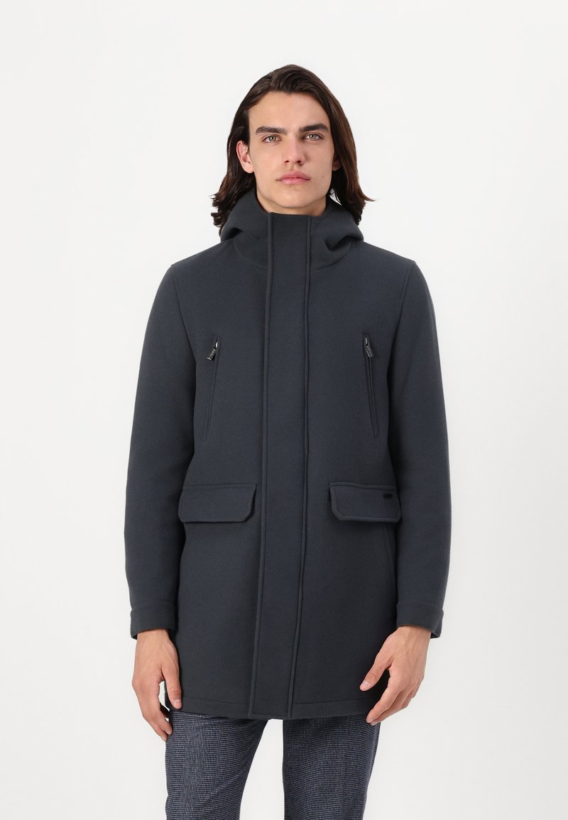 Cappotto grigio scuro con cappuccio, dotato di tasche a zip sul petto e tasche a patta, realizzato in un materiale liscio con un design strutturato. Semplice e funzionale.