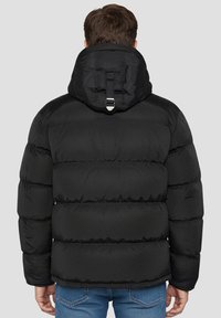 Sort dunjakke med hætte, med quiltet design, justerbar strop ved halsen og elastiske manchetter. Bruges med blå jeans.