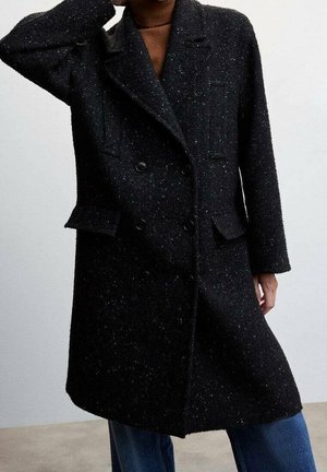 Manteau noir en laine à pois avec double boutonnage et larges revers, porté avec un jean bleu, visible des épaules jusqu'aux genoux sur un fond gris.