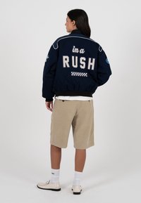 Femme aux longs cheveux foncés portant une veste bleu marine avec le texte « in a RUSH » dans le dos, un short beige longueur genou, des chaussettes blanches et des mocassins crème.