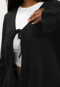 Musta neulottu neuletakki, jossa on V-pääntie, etuosassa sidottava suljenta ja ribattu hihansuut. Kangas on pehmeä ja tekstuuriltaan mielenkiintoinen.