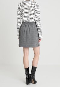 Mujer vestida con una blusa blanca con lunares, falda de cuadros en blanco y negro de cintura alta, y botas de charol negras hasta el tobillo, de pie mirando hacia atrás.