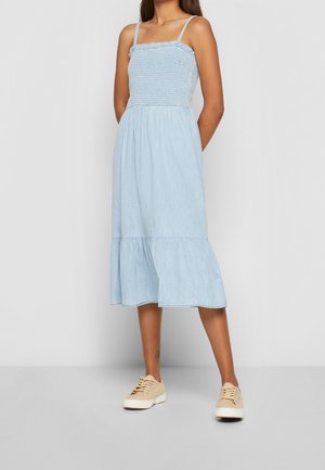 Jasnoniebieska sukienka dżinsowa z marszczonym gorsetem, cienkimi ramiączkami i falbanami u dołu. Noszona z beżowymi trampkami.