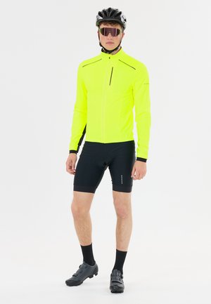 Neon-keltainen pyöräilytakki, jossa on vetoketjullinen tasku, yhdistetty mustiin shortsihin ja mustiin pyöräilykengiin. Mukana myös kypärä ja sävytetyt aurinkolasit.