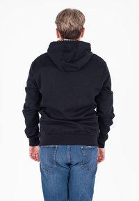 Felpa nera con cappuccio a coulisse e tasca a marsupio, abbinata a jeans di denim blu. Il tessuto sembra morbido e strutturato.