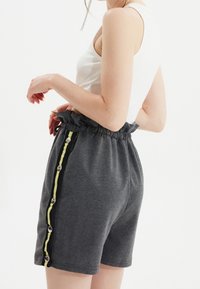 Pantaloni corti grigi con vita elastica, strisce laterali gialle e nere, e bottoni a pressione argentati lungo la cucitura sinistra. Tessuto morbido.