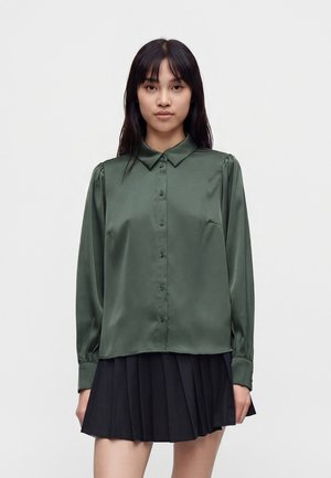 Ung kvinde med glat sort hår iført en mørkegrøn bluse med knapper og et sort plisseret skørt mod en ensfarvet baggrund.