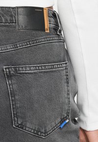 Poche arrière d’un jean en denim gris avec une étiquette de marque noire et beige et la main d’une personne reposant à proximité dans un haut blanc à manches longues côtelé.